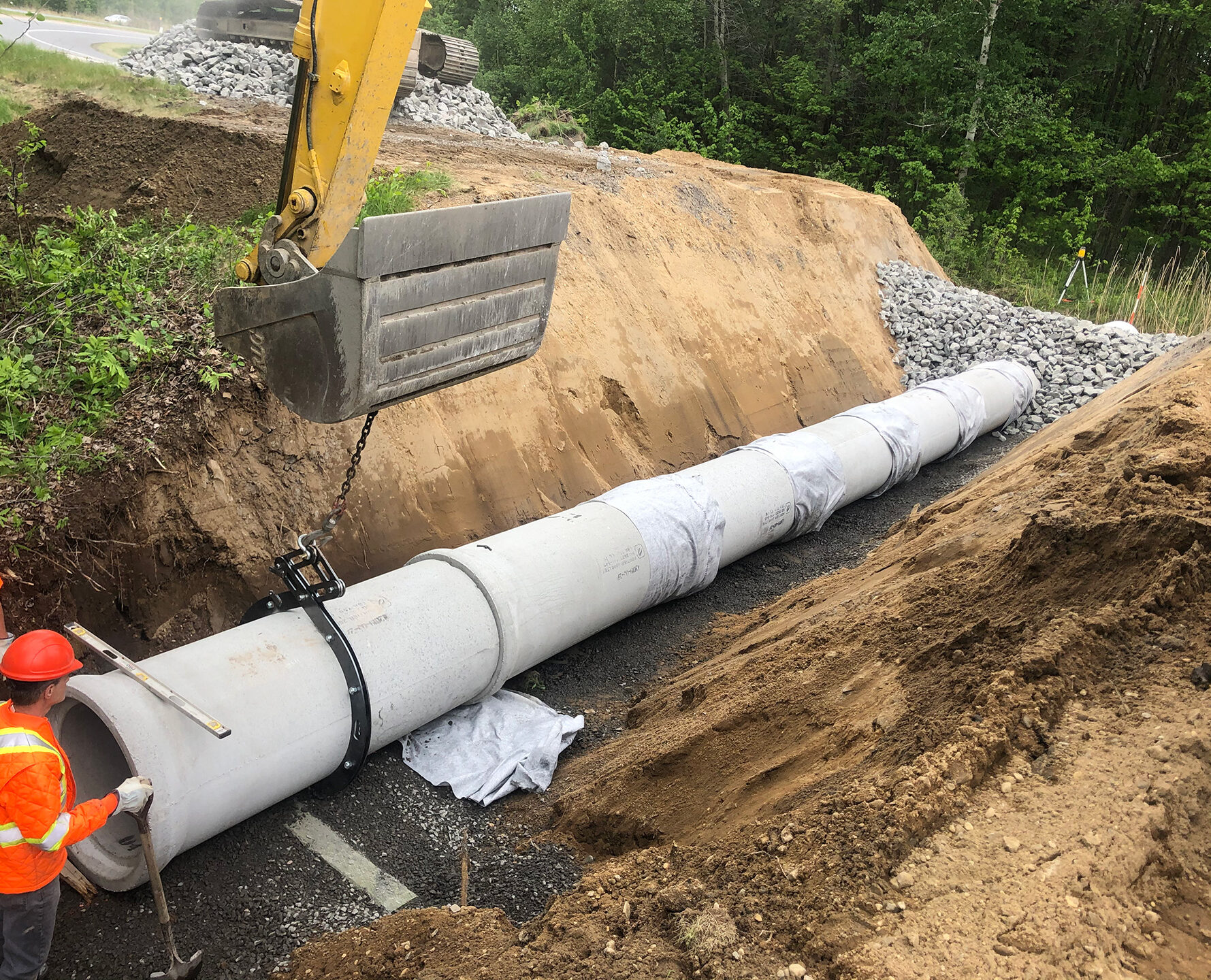 Pose d'une section de ponceau en béton dans une tranchée sur lit de gravier, guidée par un travailleur au niveau