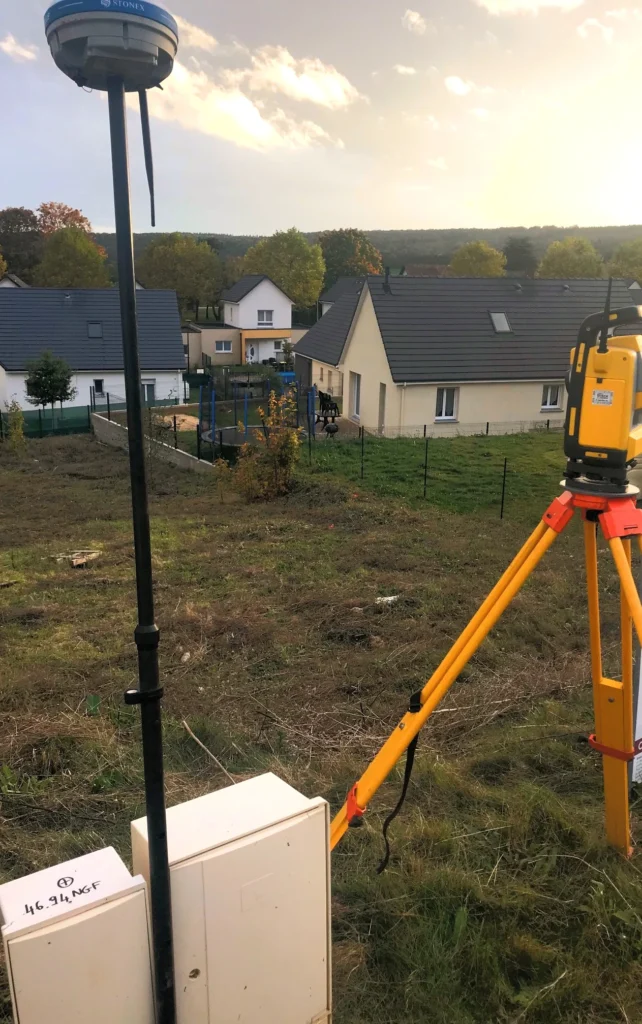 Récepteur GNSS et station totale sur trépied lors d'un relevé topographique sur un terrain surplombant des maisons